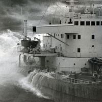 Black and white photo of ship with crashing waves.