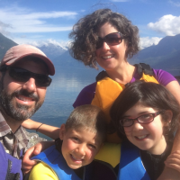 A man, a woman, and two kids all wearing life vests with a body of water and a mountain landscape in the background.