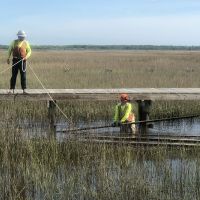 Two people in hard hats and work gear in a marsh, one standing in the water below and the other standing on a boardwalk.