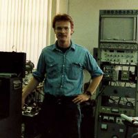 Man standing with computer equipment in the background.