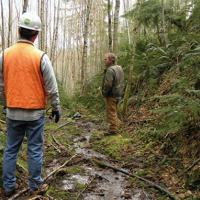 Two men standing in a wooded area. 