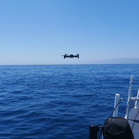 A small commercial off-the-shelf drone flying above water next to a vessel.