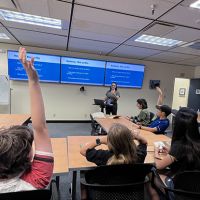 Students raise their hands to participate in a classroom setting activity led by a facilitator at the front of the classroom.
