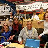 Staff and student scientists smile for a group photo in front of two banners that read 'Coastal Response Research Center at the University of New Hampshire' and 'MacFarlane Flume'.