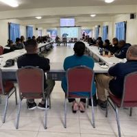 Workshop participants sit around a table in a room listening to a presenter.